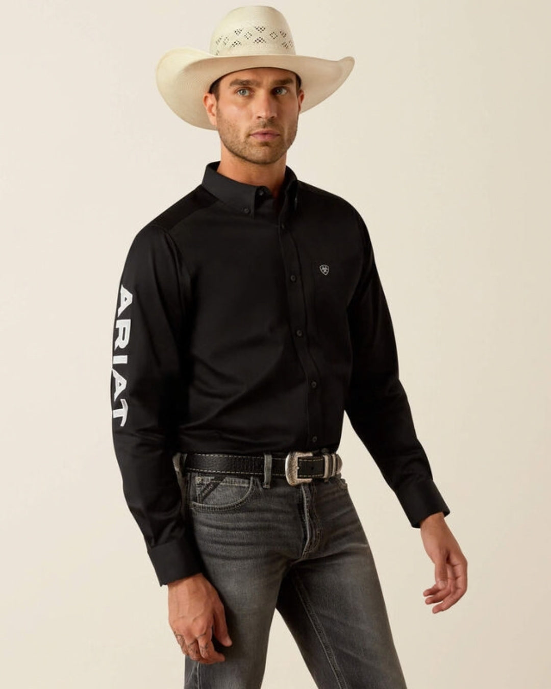Man wearing the Ariat Team Logo black twill fitted shirt with white Ariat sleeve logo, grey jeans and a straw cowboy hat
