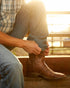 Man in jeans and a plaid shirt pulling on brown Ariat Rambler Patriot cowboy boots